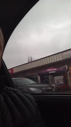 A man is sitting in the passenger seat of a moving car. He is looking at the camera. He is wearing a black quilted jacket. Outside the car window, a street scene passes by, including shops, buildings, and parked cars. The sky is overcast.