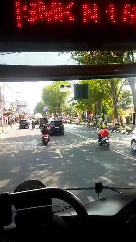 The video is shot from the perspective of someone inside a vehicle, likely a bus, looking forward through the windshield. A digital sign displaying red text, partially obscured and difficult to read, is visible at the top of the frame. The street ahead is paved and appears to be a typical urban or suburban road with trees lining the sides. Several vehicles are in motion: a dark SUV in the distance, several motorcycles, and a white car on the right side of the road. One person on a scooter, wearing a yellow helmet, is prominently featured in the right lane. The environment appears to be a sunny day, with shadows cast on the road. The scene suggests a normal flow of traffic in a populated area.