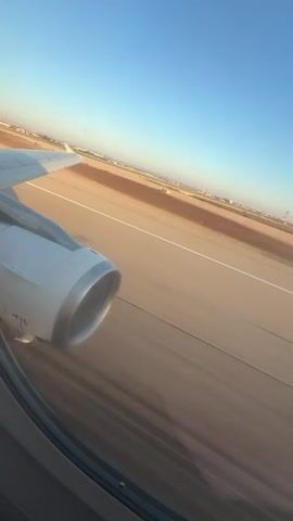 This is a first-person perspective video taken from inside an airplane window. The view is angled downwards and to the right, showing a portion of the airplane's wing and engine. The airplane is on a runway, with the ground appearing as a brown surface with white and yellow markings. In the distance, a flat landscape is visible with some structures. The sky is clear and blue. The video appears to capture the moment of takeoff or landing as the runway is moving rapidly in the frame. The overall lighting suggests it is either sunrise or sunset.