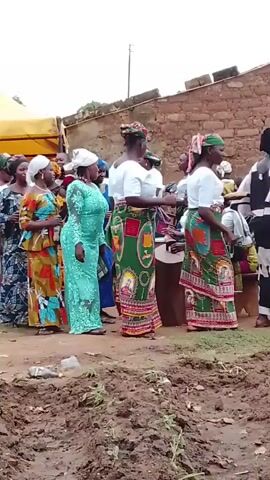 A group of women are standing and talking in an outdoor setting with dirt and construction materials visible in the foreground. The women are wearing a variety of colorful dresses and head coverings. Some of the women are holding papers. In the background, there is a yellow construction vehicle and a brick building. The overall atmosphere appears to be social. There is also music playing.