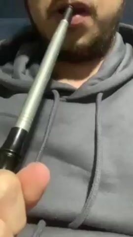 A person wearing a gray hoodie is seen holding a metal and black object to their mouth and inhaling, then exhaling a large cloud of white vapor. The person has a beard. The background is dark.