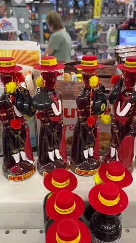 The video shows a close-up of several decorative bottles shaped like Spanish dancers. The bottles are dark red, with white and red costumes, and small red hats with yellow bands. Some bottles have small castanets in their hands. The bottles are arranged on a shelf in what appears to be a store. In the background, two people are visible, one of whom is standing behind a counter. The environment is brightly lit, with shelves of merchandise visible behind the people.