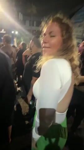A woman with blonde curly hair and fair skin is in the foreground, looking up and to her right with her eyes closed. She is wearing a white long-sleeved top with an open back and bright green pants. She has a silver chain purse slung across her body. Her left hand is on her chest. Behind her, a crowd of people are visible. One man in a black t-shirt is looking towards the woman, and another man in a red and black checkered shirt is visible further back. The scene appears to be outdoors at night, with artificial lighting creating bright spots and shadows. Buildings and tents are visible in the background.