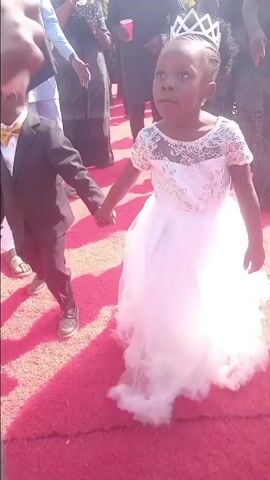 A young boy and girl are walking hand in hand on a red carpet. The boy wears a black suit with a gold bow tie, and brown shoes. The girl is wearing a white dress with lace details, and a silver crown. They are surrounded by other people, some of whom are walking, some standing. The environment appears to be outdoors. The lighting is bright and sunny. The overall atmosphere seems celebratory.
