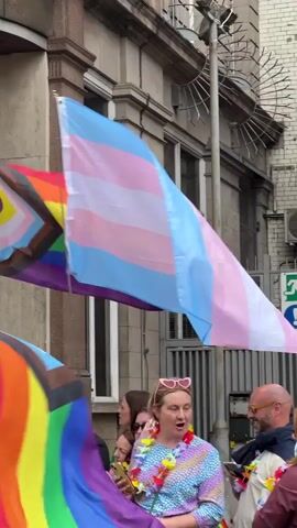 The video captures a street scene with several people holding and displaying various pride flags. In the foreground, a woman in a colorful, sequined dress and heart-shaped glasses is holding a rainbow flag. To her left, another woman with a denim jacket is also holding a rainbow flag and looking towards the camera. Behind them, a transgender pride flag, with its light blue, pink, and white stripes, is prominently displayed, waving gently. To the left of the transgender flag, a progress pride flag, which incorporates the traditional rainbow with black, brown, light blue, pink, and white stripes, is also visible. The background shows a building with windows and architectural details, and some fencing. The overall atmosphere appears festive, suggesting a pride event or parade. People are interacting with each other, and some are looking around or at the camera. The lighting suggests it is daytime, likely overcast or in shadow due to the surrounding buildings.