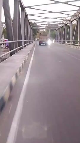 The video shows a view from a vehicle traveling across a bridge. The bridge has a metal truss structure overhead and a concrete barrier along the side. The road is paved and has a white line marking the lane. Several vehicles are visible on the bridge, including a truck, a van with red and white markings, a car, and two motorcycles. The sky appears overcast, and trees are visible on either side of the bridge. The time stamp indicates the video is from the beginning of a recording.