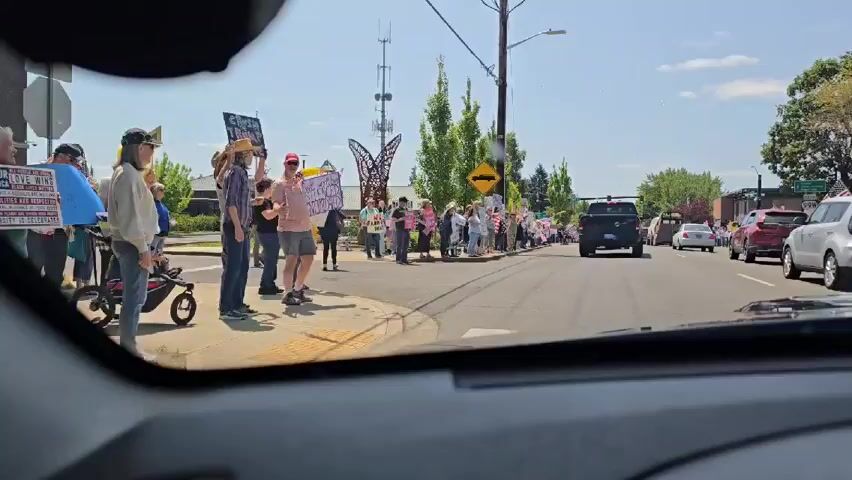 Okay, here's a concise description of the video:

The video is shot from the inside of a car, showing a street scene. A group of people are standing on the sidewalk holding signs and American flags. Some of the signs are visible, one reads "We Won't Be Silenced Intimidated", another says "No Faux-Way King", and another "This Is The People's House". Cars are driving down the street. The day appears to be sunny.