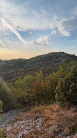 This is a vertical, handheld video of a scenic landscape under a partly cloudy sky. The sun is visible in the sky and casts a lens flare across the scene. The landscape consists of rolling hills covered in dense, green and autumnal foliage. There are visible trees in the foreground and middle ground, with some rocky terrain in the immediate foreground. The sky has wispy clouds and contrails. The video pans slightly to the right, showing more of the hilly landscape and a distant, hazy mountain range. There are no people or animals visible in the video. The overall impression is of a serene, natural environment.