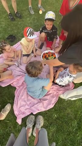 Here is a concise description of the video.
The video shows a group of about seven children sitting on a pink blanket spread out on a green lawn. They are gathered around a basket of fruit and are picking at the fruit. An adult is behind the group, assisting. Some adults are standing behind the camera view. Some of the children are wearing hats. The children look excited and are reaching for the fruit. The camera angle is directly overhead.