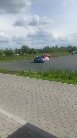The video shows a view of a race track from a paved area with a tiled walkway in the foreground. Two cars, a blue Subaru WRX STI with a large spoiler and a red hatchback, are visible on the track. The blue car is closer to the foreground and is in motion, turning on the track. The red car is further in the background, also in motion. The track is surrounded by green grass and trees. In the distance, there are some buildings and a large inflatable structure. The sky is cloudy. The video is filmed from a static position, with the camera panning slightly to follow the movement of the cars.