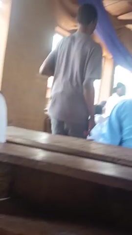 The video, likely filmed in Edu Town, Nigeria, depicts a gathering, possibly a religious service or ceremony, taking place indoors. The scene is set in a space with a corrugated metal roof supported by wooden beams. Blue and purple fabric drapes are strung across the ceiling, adding a decorative element to the rustic setting.

The primary subjects are people, mostly viewed from behind as they are seated on wooden benches. Many individuals are wearing light blue garments, suggesting a uniform or common attire for the event. In the foreground, the back of two people in blue is clearly visible, along with the edge of a wooden structure, likely part of the seating arrangement.

As the camera pans, a man in a beige shirt and grey pants enters the frame from the left, interacting with others. Further into the scene, more people are visible, including a man in a dark blue outfit and a cap, standing and speaking with others. There are glimpses of other individuals wearing patterned clothing.

The activity appears to be a formal gathering, with people seated attentively. The atmosphere seems calm and focused, consistent with a service or ceremony. The lighting suggests it is daytime, with natural light entering from openings in the structure. There is no discernible information about the weather, but the indoor setting implies protection from the elements.

A notable detail is some faint text visible on the ceiling, partially obscured by the fabric. It appears to be written in a dark color, but the content is illegible due to the quality and angle of the recording. The overall impression is a community event in a simple, functional setting.