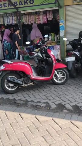 The video shows a street scene in front of a shop. Several people are visible, some of whom appear to be customers. There are also a few motorcycles parked on the street. The shop appears to be selling various items, with shelves stocked with goods visible behind the people. The ground is paved with interlocking bricks. The lighting suggests it is daytime, possibly late afternoon due to the shadows.