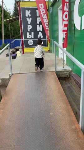 A young boy is dancing on a ramp in front of a store. He is wearing a striped shirt and black pants. The store has a green facade and a sign with Russian text. There is also a red banner with Russian text. The boy is smiling and appears to be enjoying himself.