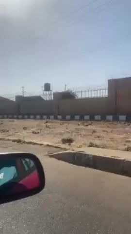 From the perspective of a moving vehicle, possibly a car, a view of a neighborhood unfolds on a clear, bright day. The architecture of the houses is visible, along with walls topped with razor wire, suggesting a focus on security. Political posters are affixed to one of the walls. Across the street, a man walks along a dirt path, near a structure that appears to be a bus stop or resting area. Power lines are visible overhead. The overall setting seems to be a developing or suburban area, possibly in a warm, dry climate.