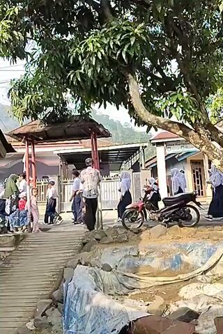 The video shows a group of students wearing school uniforms, some with hijabs, walking in front of a building. A red motorcycle is parked on the side. There are trees with lush green leaves and a hilly landscape in the background. A man wearing a patterned shirt and sarong is also present. The scene appears to be outdoors, possibly near a school or a community area.