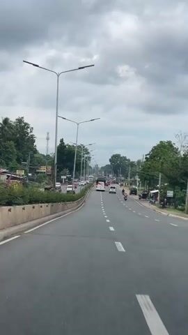 The video depicts a road with several lanes of traffic, filmed from a moving vehicle. Cars, motorcycles, and a bus are visible, moving in the same direction. Streetlights line the road. Trees and other greenery are present on either side. The sky is overcast with heavy clouds. There is concrete landscaping on the left and a commercial area on the right.