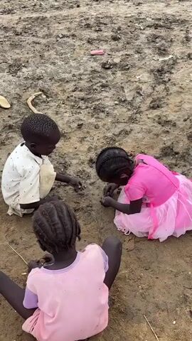 This media file depicts a scene of children playing in what appears to be a refugee camp or a similar temporary settlement. The ground is muddy and uneven, with scattered debris and some makeshift shelters visible in the background. Three young children are the central subjects, engaged in play on the ground. One child, a boy wearing a white shirt, is seated and appears to be interacting with something on the ground. Another child, a girl in a pink dress with a tiered skirt, is also on the ground, her head bowed as she plays. A third child, with braided hair and wearing a pink and purple shirt, is seated in the foreground, also focused on the ground.

The activity is clearly children playing, possibly with dirt or small objects, showcasing a moment of childhood amidst challenging surroundings. The overall setting is one of hardship, suggested by the rough terrain, the presence of basic shelters, and the general state of the environment. The lighting suggests it is daytime, with overcast or diffused light, implying no direct harsh sunlight. The weather appears to be damp, given the muddy conditions.

Notable details include the simplicity of the children's clothing and the starkness of the environment. The presence of what looks like bundled materials and tarpaulin on the shelters suggests a resource-scarce setting. The children's focus on their play, despite the surroundings, might indicate resilience or an attempt to find normalcy. There is no visible text in the media file. The location context provided is Zela, Ethiopia, which aligns with the visual cues of a potentially arid or semi-arid region experiencing difficult living conditions.