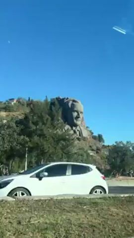 Here is a concise description of the video:

The video shows a car driving along a road.  On a hillside, there is a large sculpture of a man's head. The sky is clear and blue.