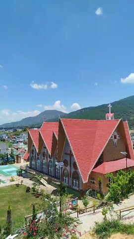 A vertical, handheld video captures a panoramic view of a village nestled between rolling green hills under a bright blue sky with scattered white clouds. The foreground is dominated by the red, gabled roofs of buildings, including a large church with intricate decorations and a cross atop its spire. To the left, a swimming pool with a bright blue surface and several submerged slides is visible. Beyond the church and pool, a collection of houses and smaller buildings forms the village, with rice paddies or green fields visible further in the distance. The video pans slightly to the right, revealing more of the lush, mountainous landscape. The overall impression is of a peaceful, sunny day in a rural setting.