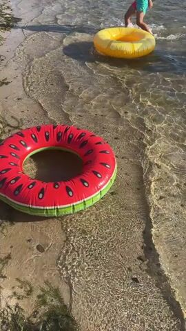 A child is running into the water at a beach. The child is wearing blue shorts. They are running towards an orange inflatable ring. Another child is in the water further out, wearing a yellow inflatable ring. In the foreground of the video is a large inflatable ring shaped like a slice of watermelon. The water is clear and shallow near the shore.