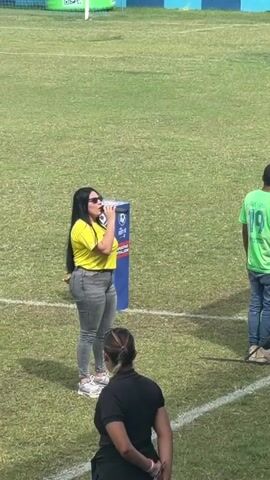 A video shows a speaker addressing an audience from a stage while people gather in the background, including a person holding a sign and another standing by a barrier. The speaker, wearing a yellow shirt and grey pants, holds a microphone and gestures with her hands as she speaks. The audience, comprising a diverse group of individuals, stands on bleachers and watches the speaker. The environment is an outdoor stadium with a grassy field, bleachers, and advertising signs. The weather appears sunny.