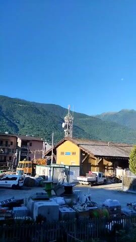 The video shows a wide shot of a rural or industrial area with mountains in the background. The sky is clear and blue. In the foreground, there is a pile of industrial materials, including what appear to be concrete blocks and spools of wire. There is a fence in front of the materials. Behind the materials, there is a yellow building with a corrugated roof, and next to it, a smaller white structure. A cellular tower is visible behind the yellow building. A white pickup truck is parked in front of the yellow building. To the left of the yellow building, there is a building with brown walls and a white truck. Further to the left, there is a yellow construction vehicle. Two green trees are visible to the right of the yellow building. In the distance, there are more buildings and mountains. There are no people visible in the video. The video appears to be taken during the daytime.