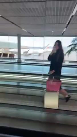 A woman is standing on a moving walkway in an airport. She is taking a selfie with her phone. She has a suitcase and a pink handbag. There is a large window behind her with an airplane visible in the background. She is dressed in a black coat and pink pants. The airport terminal appears modern and well-lit.