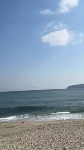 The video shows a sandy beach on a sunny day, with the ocean in the background. The sky is blue with a few small clouds. Gentle waves break on the shore. In the distance, a large ship is visible.