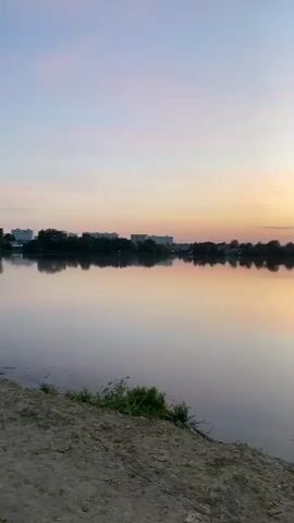 The video shows a time-lapse of a sunset over a calm body of water, likely a lake. The sky transitions from pale blues and pinks to vibrant oranges and reds as the sun sets behind the horizon. The water acts as a mirror, reflecting the colors of the sky and the silhouettes of trees and buildings along the shoreline. In the foreground, there is a dirt bank with scattered patches of green vegetation. Birds occasionally fly across the water's surface.