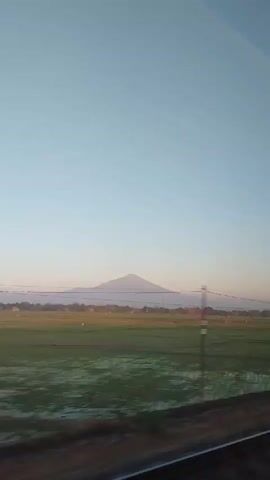 The video shows a view from a moving vehicle, likely a train, looking out at a rural landscape. In the distance, a large mountain with a conical peak dominates the horizon under a clear, pale blue sky. The foreground features a vast, green field, possibly rice paddies, with some areas appearing flooded. A power line runs horizontally across the middle ground, and a few utility poles are visible. The bottom portion of the frame shows a dark, textured surface, which is likely the window or a part of the vehicle. The lighting suggests it is either early morning or late afternoon. The overall scene is serene and natural.