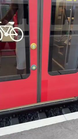 A person wearing shorts and dark socks with black clogs is seen approaching a train with red doors. A white bicycle symbol is visible on the left door. Two buttons are present on the doors, one yellow with a green circle and another blue with a green circle. The train door opens as the person approaches, revealing a step and the interior of the train. The person then steps onto the train.