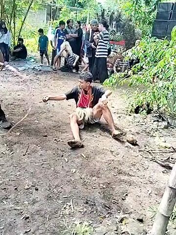 A man sits on the muddy ground with his legs splayed out, while another man stands near him holding a long, thin rope. The seated man is wearing a dark shirt and light shorts. The standing man is wearing a dark t-shirt, dark pants, and a green cap. Behind them, a crowd of people watches. A speaker is visible on the right side of the frame, along with trees and vegetation. The seated man touches the ground in front of him. There is music playing in the background.