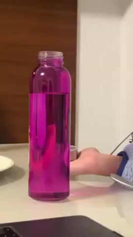 A static shot presents a clear glass bottle filled with pink liquid, standing upright on a glossy, light-colored surface. To the left of the bottle, a small white plate is partially visible. Behind the bottle, a dark brown wooden wall panel extends upwards. To the right, a white wall is visible, along with a person’s arm and hand wearing a light blue and purple textured long-sleeved garment. The person’s hand is positioned near the bottle, resting on the surface. A black laptop sits in the lower left corner. Electrical cords and a microphone are visible in the background.