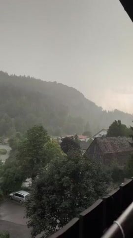 The video captures a panoramic view of a rural landscape during a heavy downpour. The foreground is dominated by a railing, suggesting the viewpoint is from a balcony or elevated structure. Below, a street is visible with a silver car parked on the left. Trees, some dense and green, others showing autumnal colors, punctuate the scene. A cluster of houses with red roofs is nestled among the trees. In the background, a lush green forest covers a misty mountain that stretches across the horizon. The sky is overcast and gray, with rain visibly falling, creating a hazy atmosphere. The overall mood is somber and peaceful, with the sound of rain creating a natural soundtrack.