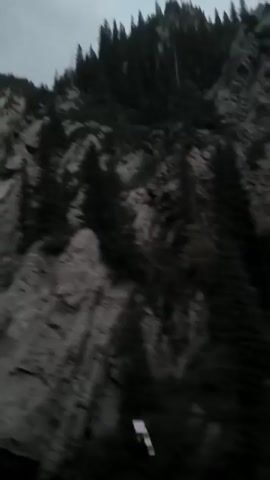 This is a shaky, first-person perspective video of a mountainous landscape. The camera is moving, likely from a vehicle, as evidenced by the blurred motion and the presence of a guardrail in the foreground. The mountainside is steep and rocky, with scattered evergreen trees growing on it. The sky is overcast and a dim light suggests it is either early morning or late evening. There are no people or animals visible in the video. The overall impression is one of a scenic, but somewhat rugged and isolated, natural environment.