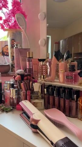The video shows a vanity table filled with various beauty products. There are numerous perfume bottles, makeup items like lipstick and foundation, a hair straightener, and a hairbrush. A framed picture of a person is visible in the background. The overall aesthetic is pink and feminine, with bright lighting. The camera pans across the vanity, showcasing the collection of items. There are no people present in the frame.