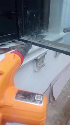 A close-up shot shows an orange heat gun being used near a window frame. A putty knife is also visible on the white windowsill. The heat gun is pointed towards the area where the putty knife is located, suggesting the removal of some material from the window frame. The background shows a dark window interior and part of a tiled wall. The area around the window appears to be under renovation or repair, with some cardboard pieces on the ground.