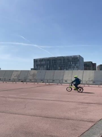 In a daytime scene under a bright, clear blue sky with a few wispy clouds and contrails, two individuals are seen riding bicycles across a wide, reddish-brown paved area. The person in the foreground, an adult, wears a neon yellow jacket and dark pants, riding a black bicycle. Behind them, a second person, appearing smaller (possibly a child), wears a blue jacket and a bright yellow helmet, riding a smaller, neon green bicycle.

They are cycling from the right side of the frame towards the left. In the mid-ground, a long, tall concrete barrier separates the paved area from several modern buildings in the background. One prominent building is tall with many windows and a distinctive architectural pattern. Other buildings are visible further in the distance. A series of low metal fences line the base of the concrete barrier. The overall atmosphere suggests a casual outdoor activity on a sunny day in an urban or semi-urban environment.