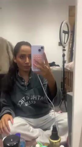 A woman is sitting cross-legged on the floor, taking a selfie in a mirror. She is wearing a gray sweatshirt with the words "ATHLETICS CLUB" printed on it and white sweatpants. She has dark hair pulled back in a low ponytail and is wearing pink lipstick. She is holding a light purple phone with a pink and white bubble-like design on the case. A white charging cable is plugged into the phone. Behind her, a ring light on a tripod is visible. Various cosmetic bottles are scattered on the floor in front of her. A fur coat hangs on the side and some items are piled up on a table or shelf in the background.
