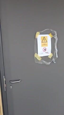 A security camera captures a gray door with a metal handle. A yellow and white warning sign with a lightning bolt symbol and a person icon is taped to the door. To the left of the door is a wall covered in vertical wooden slats. The camera pans to the right, showing more of the wooden slat wall. The camera continues to pan right, revealing the curved wall of wooden slats and a glimpse of a white ceiling and a gray carpeted floor on the left side of the frame.