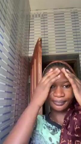 The video features a woman with dark skin and dark hair slicked back with a brown and tan scarf standing in a bathroom. She is wearing a green and white patterned shirt and has dark lipstick. She is looking directly at the camera while massaging her face with her hands in circular motions. The bathroom has a gray and white striped tiled wall and a brown door frame in the background. The lighting appears bright and natural.