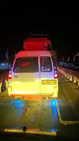 The video is a first-person perspective recording from inside a vehicle at night. The vehicle is on a flat surface, likely a ferry or a transporter, indicated by the repetitive yellow and blue lights along the surface and the structure it is on. In front of the vehicle, a white van with a red roof rack is visible. The van has license plate number B 1437 FA. The van has several stickers on its rear window and some red lettering that appears to be "DP". There are also several Indonesian flags displayed along the side of the structure the vehicles are on, with red and white colors. The background is dark, indicating it is nighttime. The overall scene suggests vehicles being transported on a ferry or similar transport across water at night.