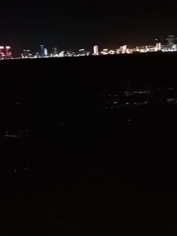 The video displays a nighttime scene over a body of water, with a city skyline in the distance. The city lights are visible as a long horizontal band of illumination across the horizon, with individual buildings and streetlights creating points of light. The water in the foreground is dark and appears to have some subtle reflections or ripples. The overall impression is of a calm, quiet night. The time of day is clearly night, indicated by the darkness and the illuminated city. There are no visible people or specific objects in the foreground, suggesting the camera is positioned some distance away, possibly from a boat or a shoreline looking across the water. The setting is coastal, given the presence of a city skyline adjacent to a large body of water, and the context provided indicates Karşıyaka, Turkey. No weather conditions are discernible, other than it is dark. There is no text visible in the video.