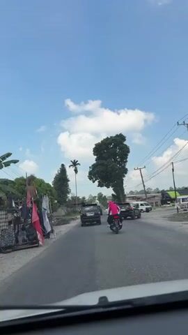 This is a vertical, first-person perspective video taken from inside a vehicle looking out through the windshield. The vehicle is moving on a paved road. In front, a motorcycle with a rider wearing a pink jacket and a helmet is traveling in the same direction. Several cars are also on the road, some in front of and beside the motorcycle. On the left side of the road, there are some trees, a tall skinny palm tree, some buildings or structures with bamboo poles, and what appear to be flags or banners attached to poles. On the right side of the road, there are more trees, including a large, tall tree with dense foliage, utility poles with wires, and some buildings. The sky is blue with scattered white clouds. The overall scene appears to be a rural or semi-rural road during the daytime.