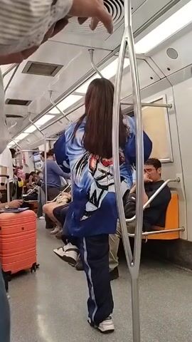 The video shows a woman dancing in the aisle of a subway car. She has long, dark hair and is wearing a blue jacket with a large anime character printed on the back, as well as dark pants with white stripes and white sneakers. She uses the vertical pole of the train as part of her movements. Passengers are visible in the background, some sitting, some standing. An orange suitcase sits on the floor near the camera. The train car has metal poles, white walls, and orange seats. The video is shot by someone standing near the pole, holding on for balance.
