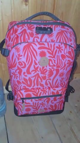 The video shows a close-up, static shot of a pink and red floral patterned backpack. The backpack is upright and facing the camera. It has a black handle at the top and black straps on the sides. There is a TSA-approved lock attached to the zippers at the top of the backpack. A circular brown logo patch with a house and trees is visible on the front. A zippered pocket is located on the lower front of the backpack. The background consists of a wooden wall and a wooden floor.