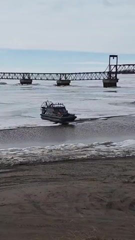 The video shows a black airboat moving through icy water near a sandy beach. A large steel bridge spans the background, connecting land on both sides of the partially frozen river. The boat has an enclosed cabin and an overhead propeller. A person can be seen standing in the boat. The sky is overcast with patches of blue. The foreground consists of a sandy area with sparse vegetation.