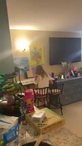 A girl with long blonde hair plays a keyboard in a living room. She is sitting on a stool facing the keyboard, with sheet music in front of her. There is a television on the wall behind her, along with two paintings and a console table. To the left of the keyboard is a fruit basket with limes and other fruits. In the foreground, there is a countertop with various items, including a box labeled "PNEUMATIC DECO NAILER," a container of garlic, and a skillet of cooked mushrooms. The lighting is warm and the environment appears to be a home.