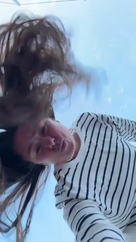 A low-angle, wide shot shows a person with long brown hair against a bright blue sky. The person is wearing a white long-sleeved shirt with black stripes and white pants. They are facing the camera and appear to be moving, with their hair flying around their head. The shot is tilted, suggesting movement or an unusual perspective.