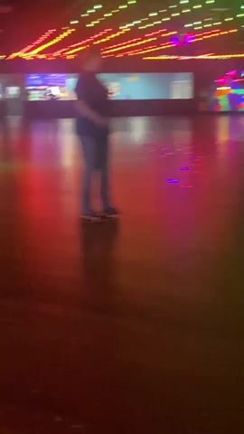 The video shows a roller skating rink with several people on roller skates. The rink is dimly lit with colorful lights reflecting off the floor. In the foreground, a person on roller skates is visible from the waist down, moving to the right. In the middle ground, a group of people are skating. On the left, two individuals are skating. In the background, there is a counter with staff and customers, and colorful murals on the wall. The lighting is dynamic, with red and blue lights casting reflections on the floor. The overall atmosphere appears to be lively and recreational.